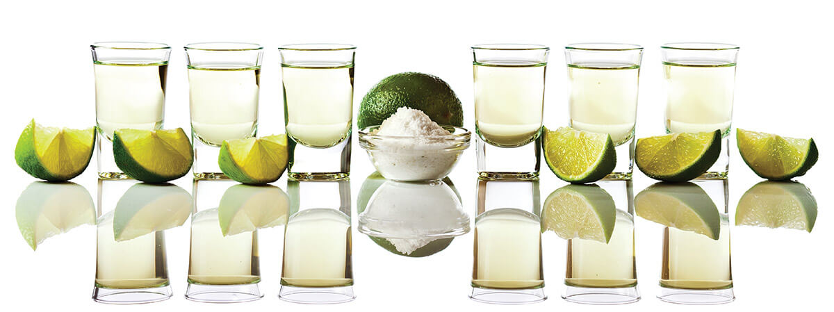Six tequila shots each with a wedge of lime. A bowl of salt and a whole lime sit in the middle.